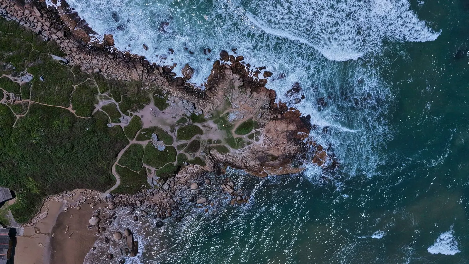 Vista aérea de cima de um costão com trilhas, rochas e ondas.